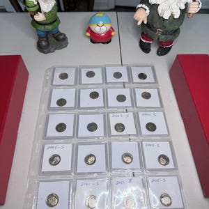 May include: A collection of silver-coloured coins housed in a clear plastic protective sheet with white cardboard holders. Several holders feature handwritten dates including 2003-S, 2000-S, 2005-S, 2008-S, 2011-S, 2013-S, and 2015-S. The coins are arranged in a grid.
