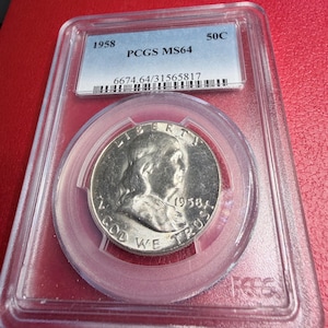 May include: A 1958 silver half-dollar coin, graded PCGS MS64, in a clear plastic holder. The coin shows a portrait and the words "Liberty" and "In God We Trust". The holder has a label with the coin's grade and serial number.