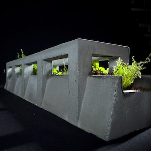 Könnte beinhalten: Ein grauer Betonblumentopf mit rechteckigen Öffnungen, in denen kleine grüne Pflanzen untergebracht sind. Der Pflanztopf hat ein geometrisches Design mit abgewinkelten Seiten und einer flachen Oberseite. Der dunkle Hintergrund hebt die Textur des Topfes und das leuchtende Grün der Pflanzen hervor.