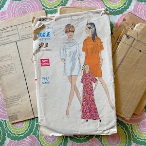 May include: A vintage Vogue sewing pattern envelope featuring illustrations of three different dress styles. The front shows a short white dress, a short orange dress, and a long patterned dress. Text includes Vogue Pattern, New Sizing, and Easy to Make.