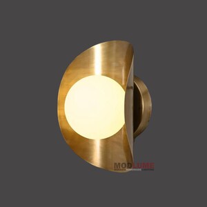 May include: A brass wall sconce with a half-moon design. The sconce features a spherical, glowing light source. The fixture has a modern aesthetic, with the text "MODLUME MODERN HANDMADE LIGHTING" at the bottom.