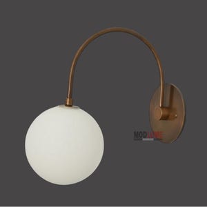 May include: A modern wall sconce featuring a spherical white glass shade and a curved, bronze-coloured arm. The sconce is mounted on a circular bronze-coloured backplate. The text "MODLUME" is visible.