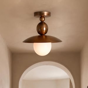 May include: A ceiling-mounted light fixture featuring a metallic bronze finish. The design includes a circular canopy, two stacked spherical elements of varying sizes, a wide flat disc-shaped shade, and a single spherical white glass globe.