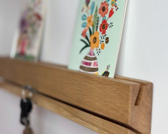 May include: A wooden wall-mounted shelf with a key rack. The shelf holds two colorful floral art cards. The keys hang below the shelf. The shelf is made of light-colored wood.