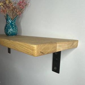 Puede incluir: Estante de madera clara con soportes metálicos negros. Un jarrón turquesa con diseño floral contiene flores secas. El conjunto crea un ambiente rústico y sencillo, ideal para decoración.