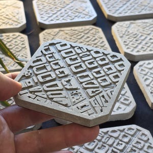 Puede incluir: Un posavasos hexagonal de color gris claro con un patrón geométrico en relieve de rectángulos y cuadrados interconectados. La superficie tiene un acabado mate y texturizado con una capa base oscura visible en el lateral.
