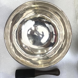 Puede incluir: Un cuenco tibetano plateado con una maza de madera oscura. El cuenco tiene una superficie lisa y reflectante y forma ovalada. La maza tiene un extremo de golpeo rectangular y un mango redondeado. Los objetos están sobre una superficie gris claro.