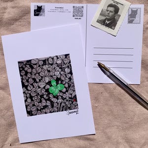 May include: A white postcard featuring a green four-leaf clover illustration on a gray background. A second postcard with a postage stamp and a pen are also visible. The artist's signature is at the bottom of the postcard.
