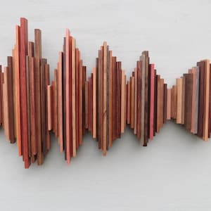 May include: A wooden wall art piece composed of vertical wooden slats in varying shades of brown, red, and pink. The arrangement creates a wave-like pattern, adding a dynamic visual element. The artwork is mounted on a light-colored wall.