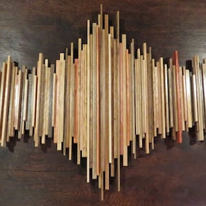 May include: Wooden wall art sculpture resembling a sound wave. The artwork is composed of vertical wooden slats in varying shades of brown, beige, and red, creating a dynamic, textured design. The piece is mounted on a dark brown wooden surface.