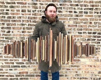 May include: A wooden wall art piece composed of vertical wooden slats in various shades of brown, gray, and beige. The artwork is held by a person in front of a brick wall. The wooden slats vary in length and width, creating a textured, abstract design.