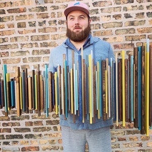 May include: A wooden wall art piece composed of vertical wooden slats in various colors, including blue, yellow, and brown. The artwork is held in front of a brick wall, showcasing a modern, abstract design. The slats vary in height, creating a dynamic visual.