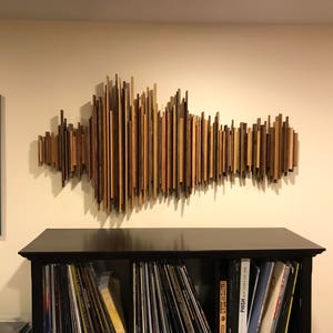 Peut inclure: Une œuvre d'art en forme d'onde sonore en bois, composée de lattes de bois de différentes nuances de brun, fixée au mur. L'œuvre est au-dessus d'un meuble noir rempli de disques vinyles. Deux œuvres encadrées sont de chaque côté.