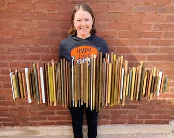 May include: A wooden wall art piece composed of various vertical wooden slats in shades of brown, white, yellow, and pink. The artwork is held in front of a brick wall. The wooden slats are of varying lengths and widths, creating a textured, abstract design.