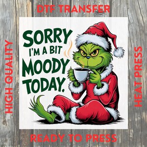 May include: A green cartoon character in a Santa suit, holding a coffee cup, with the text "Sorry I'm a bit moody today." The image is a DTF transfer, ready to press, with high quality.