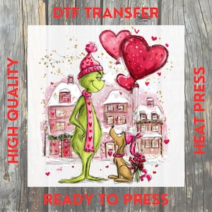 May include: A whimsical illustration featuring a green character with a pink hat and scarf, holding a heart-shaped balloon. A dog with a pink bow and flowers stands nearby. The background shows snow-covered buildings and hearts. Text reads "DTF TRANSFER", "HIGH QUALITY", and "READY TO PRESS".