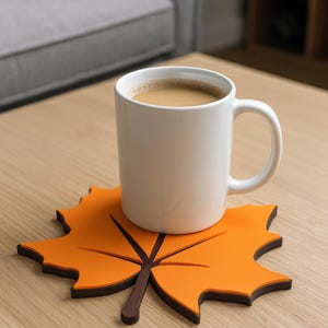 May include: A white ceramic mug filled with coffee rests on an orange leaf-shaped coaster with a brown outline and stem. The mug has a curved handle and is on a light wood surface.