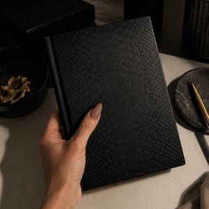 May include: A rectangular notebook featuring a black cover with a textured snakeskin pattern. The notebook has a bound spine and a consistent, repeating scale-like design across the entire front surface, providing a tactile and detailed appearance.