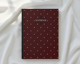 Puede incluir: Un cuaderno granate con lomo negro y lunares blancos. La palabra "NOTEBOOK" está impresa en una fuente delicada en la parte superior, con un diseño adornado encima. El cuaderno está sobre una tela blanca y sedosa.