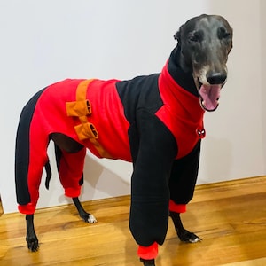 Pode incluir: Um macacão para cães preto e vermelho. O macacão é vermelho com mangas e pernas pretas. O cão é um galgo com a boca aberta. O macacão tem detalhes em laranja e um pequeno emblema no peito.