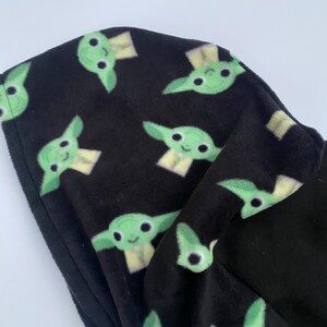 Puede incluir: Tela negra con un patr&oacute;n repetido de caras de personajes de dibujos animados verdes. El personaje tiene ojos grandes, orejas puntiagudas y una peque&ntilde;a sonrisa. La tela parece suave y afelpada.