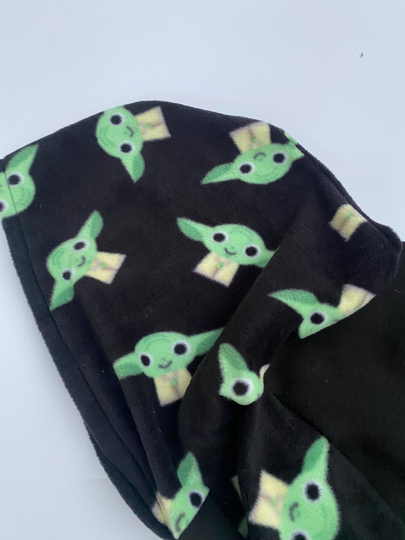 Puede incluir: Tela negra con un patr&oacute;n repetido de caras de personajes de dibujos animados verdes. El personaje tiene ojos grandes, orejas puntiagudas y una peque&ntilde;a sonrisa. La tela parece suave y afelpada.
