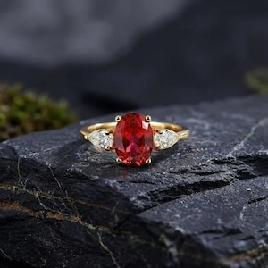 Peut inclure: Bague en or ornée d'une grande pierre rouge ovale et de deux pierres blanches en forme de poire. La bague est posée sur une surface sombre et texturée. Les couleurs des pierres précieuses sont vives.