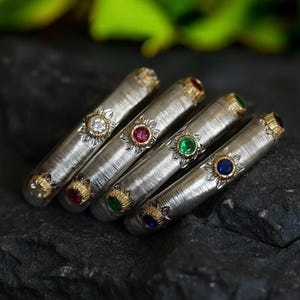 Può includere: Primo piano di quattro braccialetti argentati con dettagli dorati e pietre preziose colorate. Ogni braccialetto presenta una pietra diversa: un diamante, un rubino, uno smeraldo e uno zaffiro. I braccialetti sono disposti su una superficie scura e strutturata.