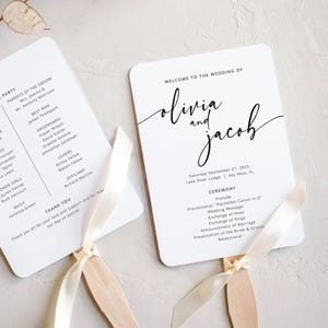 May include: White wedding fan with the text "Welcome to the Wedding of Olivia and Jacob" in elegant script. The fan includes the wedding date, location, and ceremony details. A cream-coloured ribbon is attached to the wooden handle.