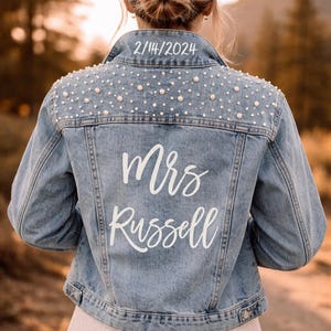 Könnte beinhalten: Hellblaue Jeansjacke mit Perlenbesatz auf den Schultern. Auf der Rückseite der Jacke steht in weißer Schrift "Mrs Russell", darüber das Datum "2/14/2024". Eine stilvolle Brautjacke.