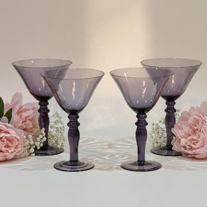 May include: Four purple glass martini glasses with wide, conical bowls and thick, rounded stems. The glasses are arranged in a row, interspersed with soft pink peony flowers and small white baby's breath sprigs, creating a balanced display.