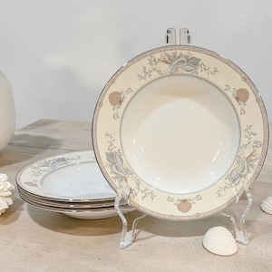 May include: A set of round ceramic soup bowls featuring a cream-colored center and a wide rim decorated with a pattern of tan and grey seashells, delicate foliage, and a thin silver metallic border. One bowl is displayed upright on a clear stand.