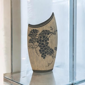 May include: A tall, tapered ceramic vase with a textured beige surface featuring a dark brown, etched graphic of a grape cluster with leaves and vines. The vase has an asymmetrical, slanted top rim and a dark brown base, creating a distinct contrast.