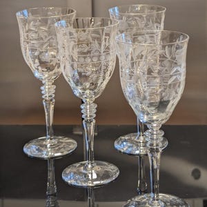 May include: A set of four clear glass goblets featuring delicate etched floral patterns and horizontal bands. Each stem is designed with a series of stacked rings leading down to a circular base, providing a consistent look across all four pieces.