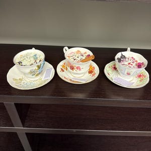 Puede incluir: Tres velas en tazas de té sobre platillos, cada una con un diseño floral único. Las velas están llenas de flores secas y especias. Una vela tiene una ramita de canela. Las tazas de té están sobre un estante marrón oscuro.