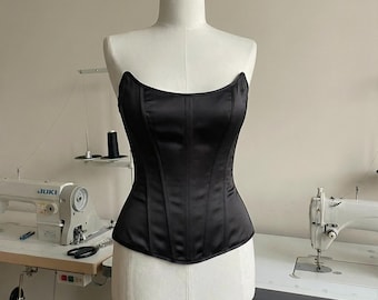 May include: A black satin corset displayed on a mannequin. The corset has a structured design with vertical seams and a curved neckline. The garment is displayed in a studio setting with sewing machines in the background.
