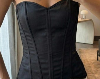 May include: A black corset with a sweetheart neckline. The structured bodice features vertical seams and a smooth, satin-like finish. The corset is paired with white pants. A silver cuff bracelet is visible on the left wrist.