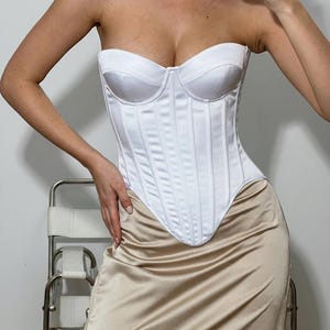 May include: A white strapless corset top with vertical paneling is paired with a champagne-colored satin skirt. The corset has a structured, bustier-style design. The outfit is a formal or evening wear ensemble.