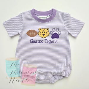 May include: A light purple and white striped baby romper with short sleeves. The romper features a football, tiger face, and paw print design. The text "Geaux Tigers" is embroidered below the design. The romper has a purple collar.