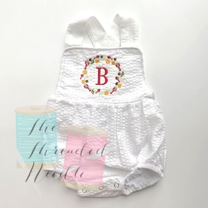 May include: White seersucker baby romper with a floral wreath and the letter "B" embroidered on the front. The romper has a bubble silhouette, adjustable straps, and snap closures at the bottom. The overall design is a classic and timeless style.