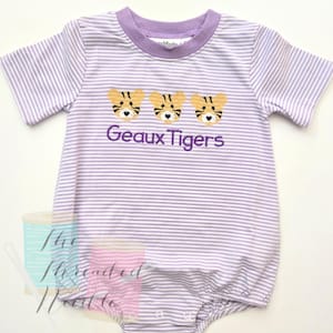 May include: A baby's romper with horizontal lavender and white stripes. The romper has a lavender collar and sleeve trim. Three embroidered tiger faces are above the text "Geaux Tigers" in purple thread.