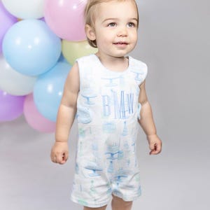 May include: A baby wearing a white romper with a blue cake and cake stand pattern. The romper has the initials "BMW" in blue. Pastel balloons in pink, blue, and purple are in the background.