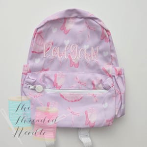 May include: A lavender backpack with a pattern of pink ballet shoes, bows, and hearts. The name "Raegan" is embroidered in a matching pink thread. The backpack has a white zipper and two side pockets.