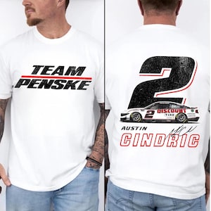 May include: White t-shirt featuring the text "TEAM PENSKE" in black with a red stripe. The back of the shirt displays a large number "2" with a race car graphic and the name "AUSTIN CINDRIC" in red.