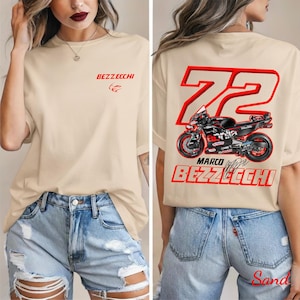 以下が含まれることがあります： 前面に赤い「BEZZECHI」の文字とオートバイのロゴが入ったベージュのTシャツ。背面には大きな赤い「72」のグラフィック、オートバイのイラスト、そして「MARCO BEZZECHI」の文字が描かれています。Tシャツはデニムショーツと合わせています。