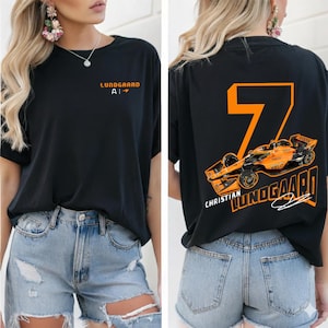 May include: Black t-shirt featuring the name "Lundgaard" and the initials "AI" in orange on the front. The back displays a large orange number "7" with a race car graphic and the name "Christian Lundgaard".