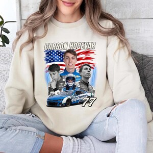 May include: A cream-colored crewneck sweatshirt featuring a graphic print of race car driver Carson Hocevar. The design includes three portraits of the driver, a stylized American flag background, and a blue number 77 race car.
