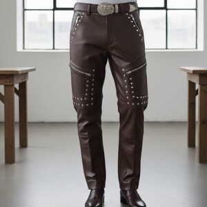 May include: Brown leather pants with silver zipper details and studded accents. The pants feature a decorative belt with a silver buckle. The pants are worn with brown leather boots. The pants are a stylish addition to any wardrobe.