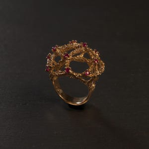 Può includere: Un anello in oro caratterizzato da una struttura organica e testurizzata simile a rami che forma una cupola. La superficie del metallo è ruvida e porosa, arricchita da piccole pietre preziose rotonde di colore rosso vivo incastonate nel design.
