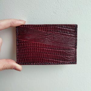 Può includere: Un porta carte bordeaux con un motivo strutturato simile a quello di un rettile. Il portafoglio rettangolare è realizzato in materiale rosso scuro e presenta cuciture nere sui bordi. Il porta carte è tenuto da una mano.